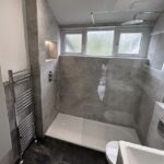 A modern bathroom design with dark floor tiles and grey wall tiles, featuring a walk-in shower with a glass partition, a white toilet, and a chrome towel radiator.