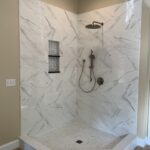 A modern corner shower with marble-like tiles and glass doors, designed for a bathroom remodel, featuring a rain shower head and built-in shelf.