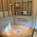 A bathroom remodel underway with exposed pipes, a shower base installed, and a partially installed usg durock cement board.