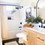 A modern bathroom design featuring white subway tiles, a wooden vanity with a black countertop, gold accents in the lighting and fixtures, and decorative plants. A patterned rug lies on the floor next to