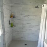 A modern bathroom interior featuring a walk-in shower with white marble-patterned tile walls, a glass shower door, and various toiletries placed on built-in shelves next to a sleek bathroom cabinet, with a