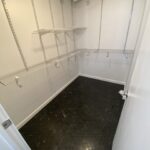 Empty walk-in bathroom remodel with white walls and several rows of metal wire shelving on three sides, featuring a gray and black hexagonal tile floor.
